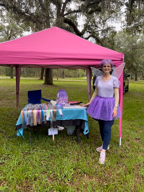 Tambra of The Hair Sparkler as a traveling fairy hair vendor at an outdoor festival, wearing fairy wings beside her tinsel table